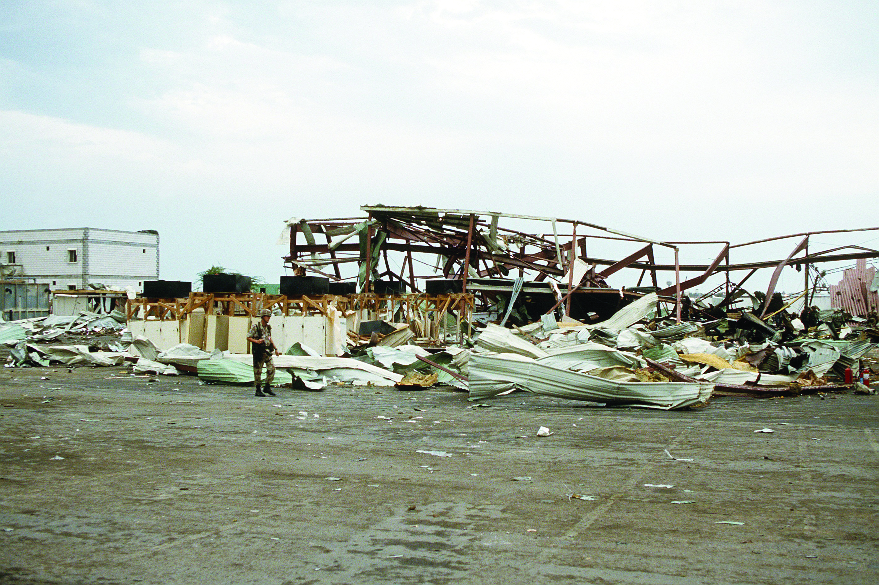 The aftermath of a Scud missile attack, showing a completely destroyed warehouse. A lone figure stands amidst the widespread debris and twisted metal structure.